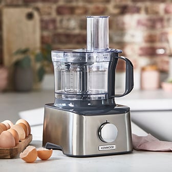 Das Bild zeigt eine silberfarbene Kenwood Küchenmaschine mit einem transparenten Behälter und Deckel auf einer hellen Arbeitsfläche in einer Küche, neben einem Holzgefäß mit braunen Eiern und zwei aufgebrochenen Eierschalen.