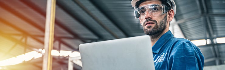 Ein Arbeiter mit weissem Schutzhelm in einer Halle mit einem Laptop in der Hand und Schutzbrillen auf.