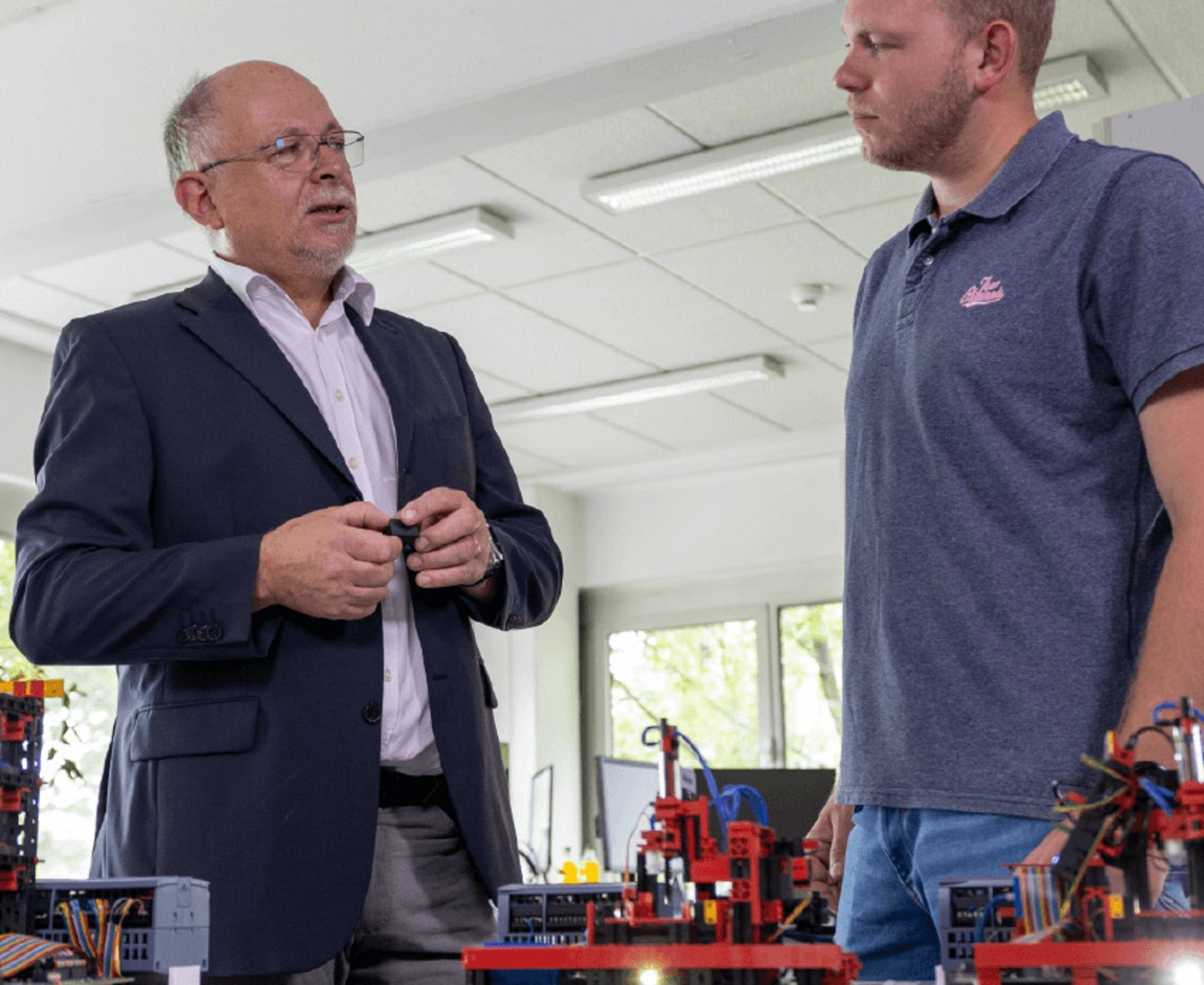 Wolfgang Lex, in einem dunklen Anzug und ein jüngerer Mann in einem grauen Poloshirt stehen sich in einem hellen Raum gegenüber und unterhalten sich über Module aus dem Agile Production System, welche im Vordergrund zu sehen sind.