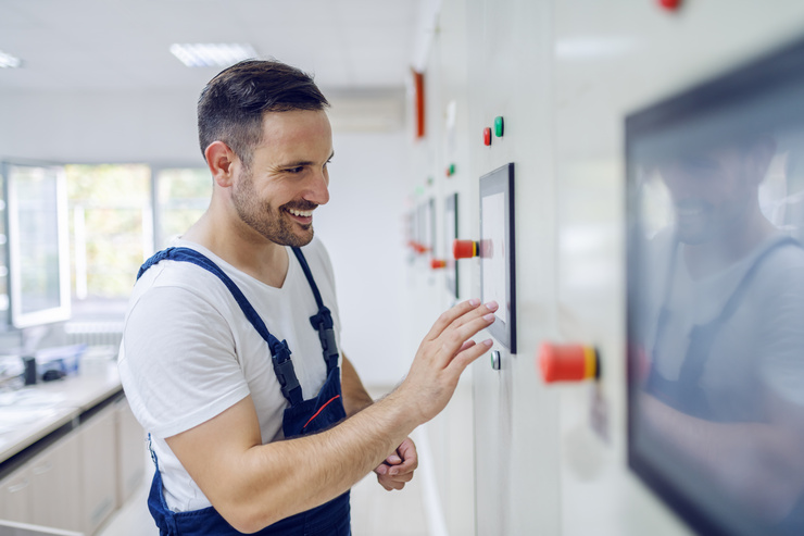Ein lächelnder Techniker in Arbeitskleidung bedient ein digitales Bedienfeld in einem hellen Technikraum.