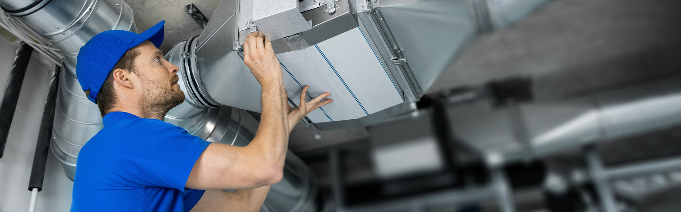Handwerker in blauer Arbeitskleidung montiert ein silbernes Lüftungsrohr an einer Betondecke.