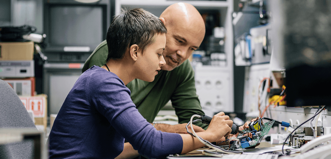 Zwei Personen, ein Erwachsener und ein junger Erwachsener, arbeiten gemeinsam an einer elektronischen Platine.