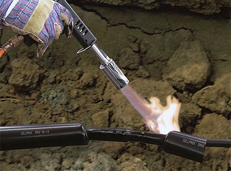 Ein Arbeiter erwärmt mit einer Gasflamme einen schwarzen Warmschrumpfschlauch über einer Kabelverbindung in einem Erdgraben, um diese wasserdicht zu versiegeln.