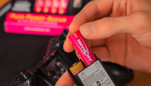 Eine Person setzt eine pinkfarbene Ansmann-Akkubatterie in das Batteriefach eines schwarzen Game-Controllers ein, während die Produktverpackung im Hintergrund unscharf zu sehen ist.
