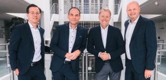 Die Geschäftsführung von Conrad Electronic, bestehend aus (von links nach rechts) Dr. Sebastian Dehnen, Ralf Bühler, Dr. Daniel Saga und Guy Dann, steht lächelnd nebeneinander an einem Geländer in einem modernen, hellen Büro-Atrium.