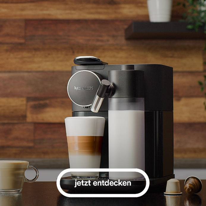 Das Bild zeigt eine schwarze Nespresso Gran Lattissima Kaffeemaschine auf einer dunklen Oberfläche vor einer holzvertäfelten Wand, mit einem hohen Glas gefüllt mit weißer Flüssigkeit und einem Glas mit geschichtetem Kaffee daneben, sowie zwei Kaffeekapseln und einer kleinen Tasse im Vordergrund und einem hervorgehobenen Button mit der Aufschrift "jetzt entdecken".