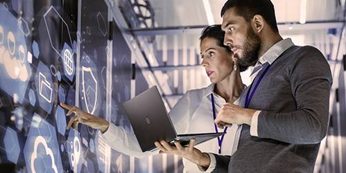 Eine Frau in einem hellen Hemd und ein Mann in einem grauen Pullover stehen in einem Raum mit Serverracks und betrachten gemeinsam eine große digitale Anzeige mit leuchtenden Symbolen, wobei die Frau mit dem Finger auf den Bildschirm zeigt und der Mann einen Laptop hält, was auf eine technische Problembehandlung und die Möglichkeit der Kontaktaufnahme mit Ansprechpartnern für persönliche Anliegen hinweist.