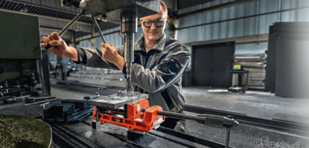 Ein Metallarbeiter mit Schutzbrille bedient eine Säulenbohrmaschine, um ein in einem orangefarbenen Schraubstock eingespanntes Werkstück in einer Werkstatt zu bearbeiten.