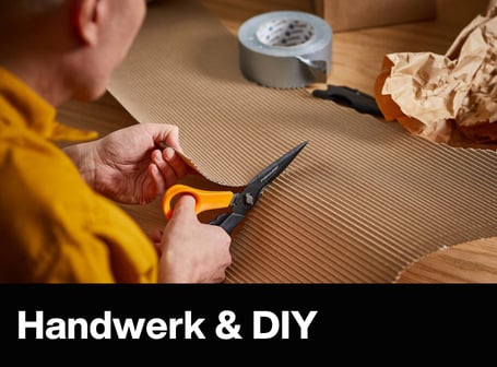 Eine Person schneidet mit einer orangefarbenen Fiskars-Schere ein Stück Wellpappe auf einem Holztisch zurecht.