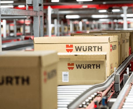 Mehrere beige Kartons mit dem roten und schwarzen Würth-Logo werden auf einer automatisierten Förderanlage in einem Logistikzentrum transportiert.