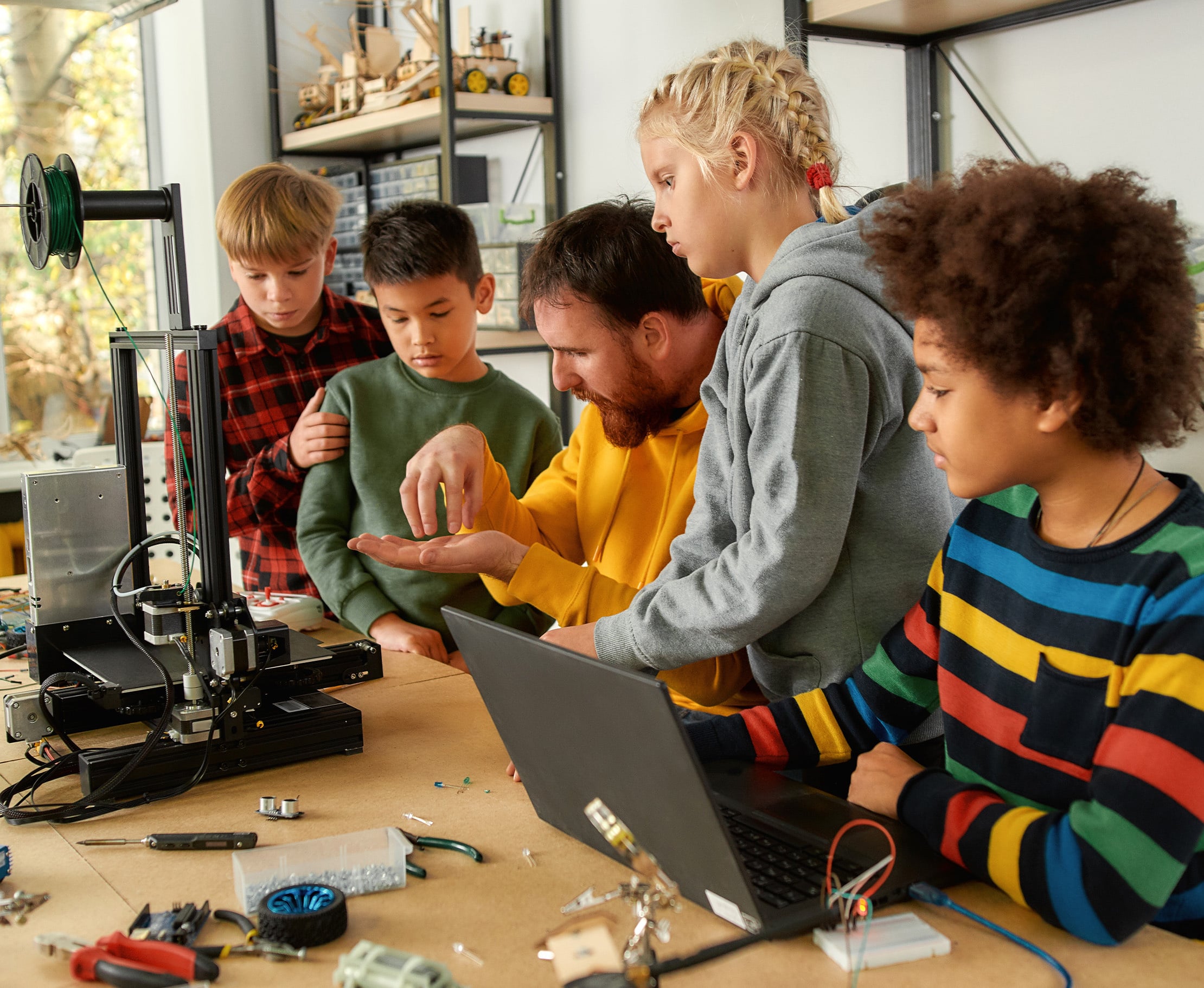 Weitwinkelaufnahme von Schülern und Lehrer im Technikunterricht mit 3D-Drucker und Laptop. Technische Modelle im Hintergrund.
