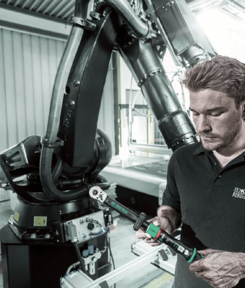 Ein Techniker hält einen Wera-Drehmomentschlüssel mit grün markierter Skala in den Händen, im Hintergrund ist ein großer schwarzer Industrieroboter in einer Werkshalle zu sehen.