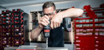 Ein Handwerker mit Schutzbrille bohrt mit einer schwarzen Akkubohrmaschine konzentriert ein Loch in ein Metallstück, während im Hintergrund Regalsysteme mit roten und schwarzen Boxen zu sehen sind.
