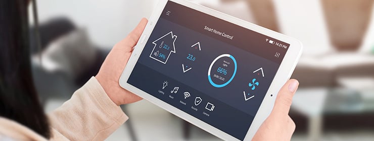 Person hält ein Tablet mit einer Smart-Home-Steuerungs-App, die Temperatur, Luftfeuchtigkeit und Luftqualität anzeigt.