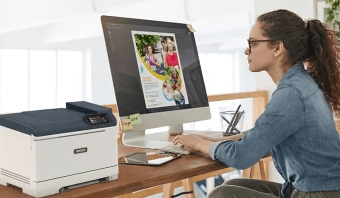 Eine junge Frau mit Brille sitzt an einem Holztisch vor einem großen Computermonitor, auf dem ein farbiges Dokument angezeigt wird, und tippt auf einer Tastatur, während links ein Xerox Drucker steht.