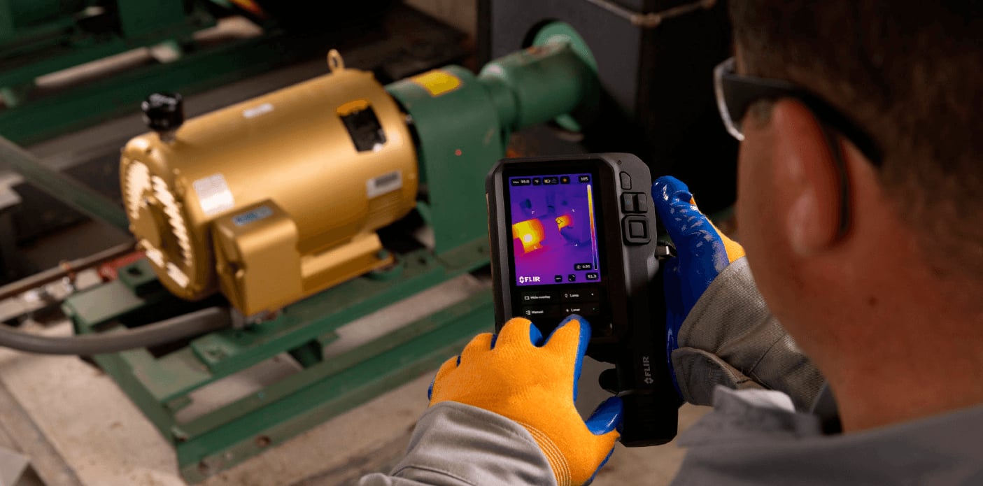 Eine Person mit Schutzhandschuhen bedient eine FLIR Wärmebildkamera, um einen goldenen Elektromotor in einer Industrieumgebung zu überprüfen.