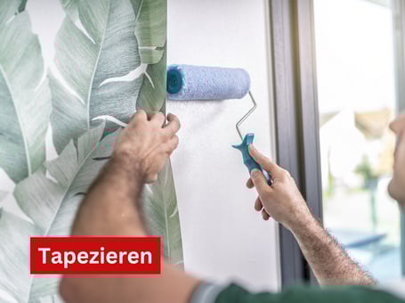 Zwei Hände halten eine Farbrolle und streichen damit Kleister auf eine weiße Wand zum Tapezieren.