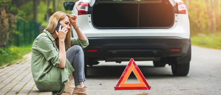 Eine Frau telefoniert neben einem liegengebliebenen Auto mit geöffnetem Kofferraum und Warndreieck auf der Straße.