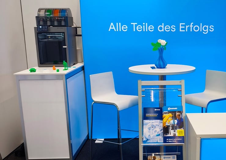 Eine Nahaufnahme des Messestandes, worauf ein Tisch mit zwei Stühlen und einem 3D Drucker zu sehen sind
