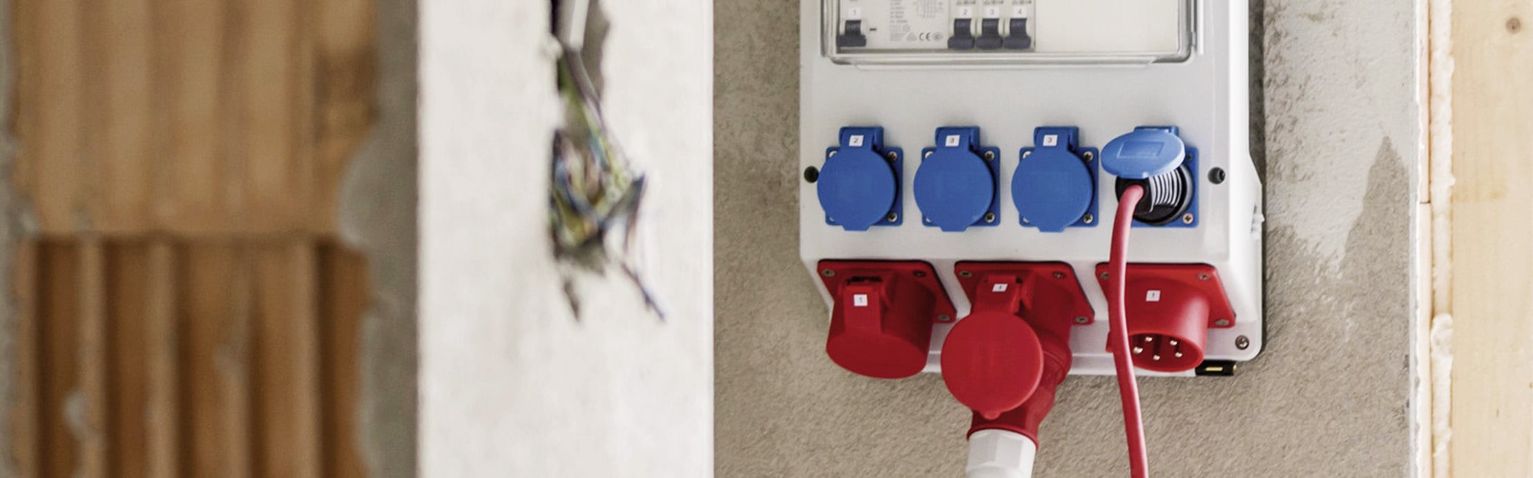 Ein weißer Baustromverteiler mit drei blauen und drei roten CEE-Steckdosen ist an einer Wand montiert, wobei ein rotes Kabel in eine blaue und eine rote Steckdose gesteckt ist.