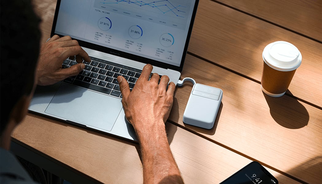 Person tippt auf einem Laptop mit Datenanzeige auf einem Holztisch. Eine weiße Powerbank ist an den Laptop angeschlossen. Daneben stehen ein Kaffeebecher und ein Smartphone.