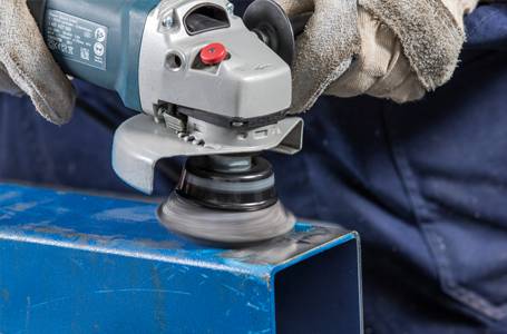 Eine Person in robusten Arbeitshandschuhen bearbeitet ein blaues Metallprofil mit einem Winkelschleifer, der mit einer rotierenden Topfbürste ausgestattet ist.