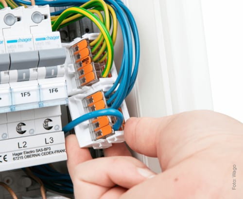 Eine Nahaufnahme einer Hand, die ein blaues Kabel an einer orangefarbenen Wago-Klemme in einem Sicherungskasten befestigt.