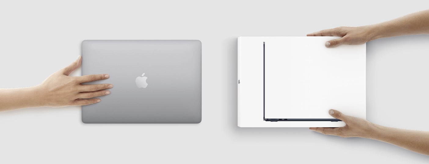  Eine Nahaufnahme von zwei Händen, die von links und rechts kommen und einen grauen Apple MacBook Laptop und ein weißes, geschlossenes Laptop, das von der Seite betrachtet wird, halten.