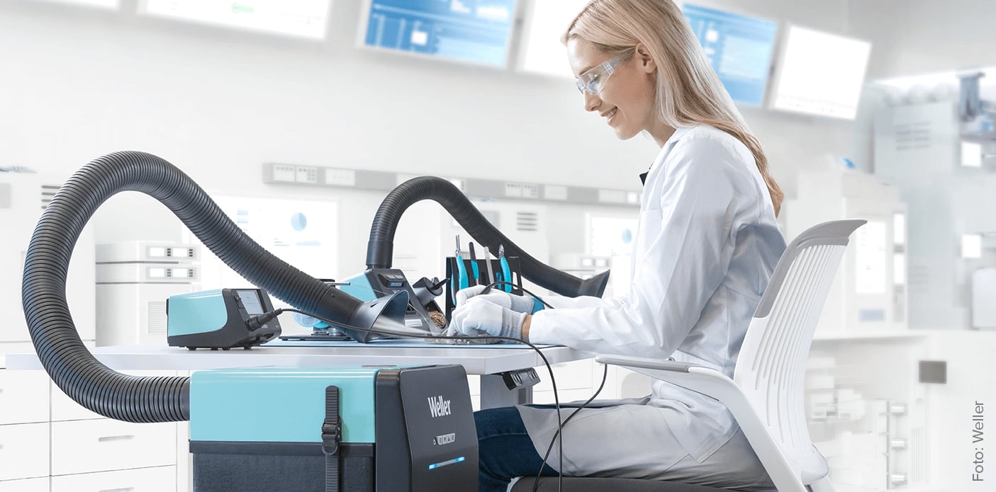 Das Bild zeigt eine Frau in einem Labor- oder Werkstattumfeld, die an einem Tisch mit elektronischen Komponenten arbeitet. Sie trägt Schutzbrille und Handschuhe, was auf Tätigkeiten im Bereich Elektrotechnik oder Elektronik hindeutet. Im Vordergrund steht ein neues, ESD-sicheres ZeroSmog Guard Absauggerät. Diese Geräte gewährleisten durch ihre effiziente Filtration von Lötrauchpartikeln und -gasen eine erstklassige Luftqualität.