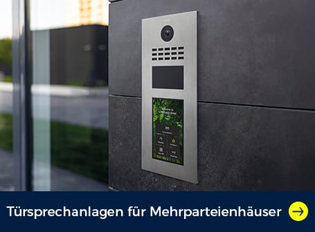 Eine moderne Türsprechanlage in Edelstahloptik mit Monitor auf einer dunklen Fassadenwand neben einer großen gläsernen Eingangstür montiert.