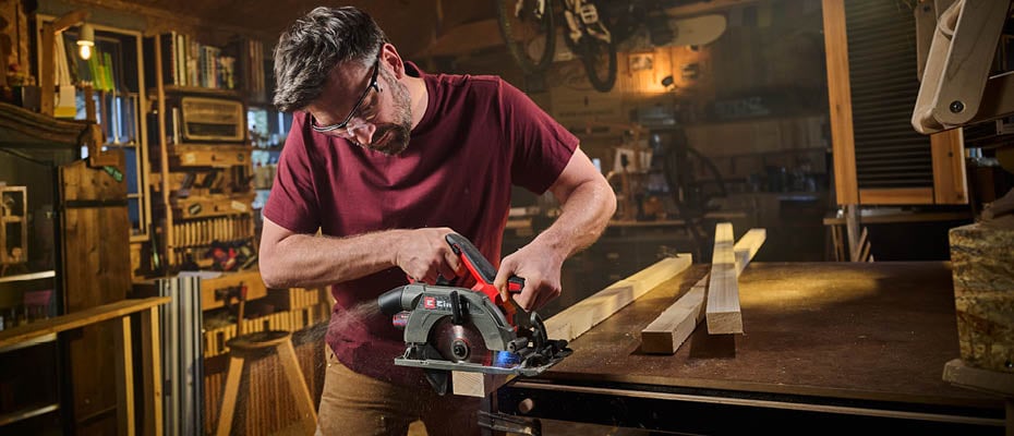  Ein bärtiger Mann mit Schutzbrille und rotem T-Shirt sägt in einer Werkstatt mit einer roten Einhell Handkreissäge ein Holzbrett.
