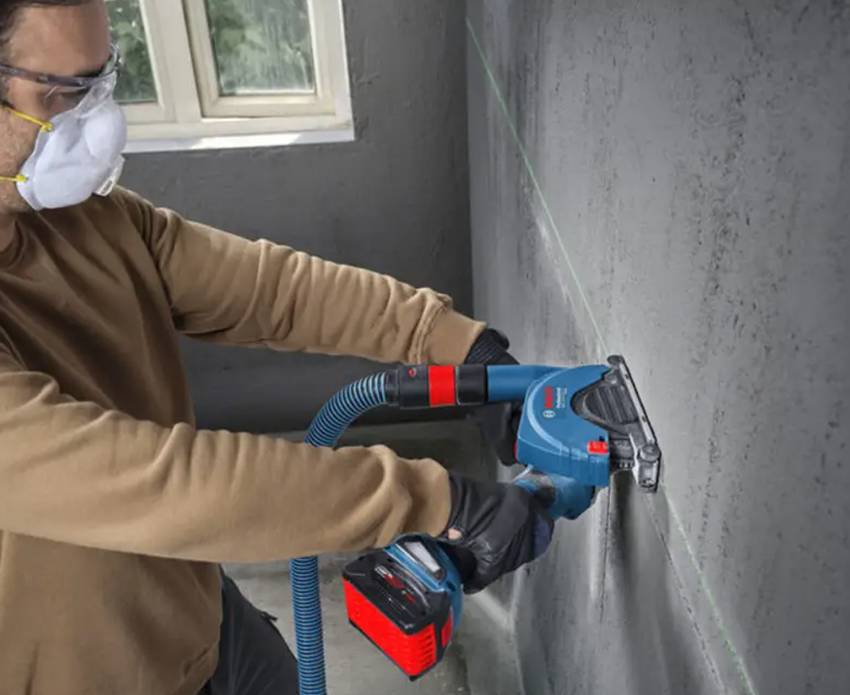 Ein Handwerker mit Mundschutz und Schutzbrille fräst Schlitze in eine Betonwand mit einem Bosch-Akku-Winkelschleifer GWS 18 V-11PS und angeschlossenem Staubabsaugschlauch.