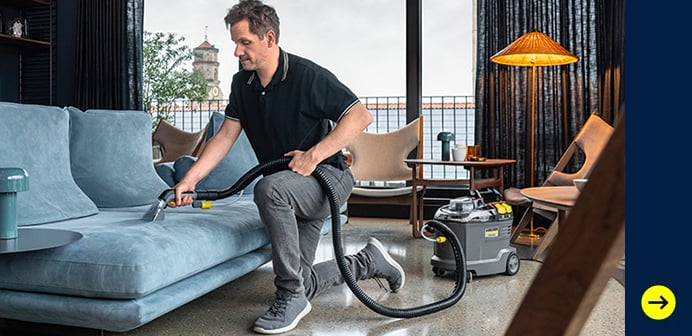Auf dem Bild erkennt man einen Mann, der mit einem Staubsauger ein graues Sofa reinigt. Im Hintergrund ist ein Wohnzimmer mit Möbeln und einer Lampe zu sehen. Am unteren Rand steht "Zu Kärcher Professional" und ein gelber Pfeil.