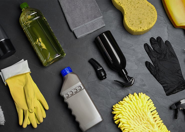 Verschiedene Reinigungsutensilien wie Handschuhe, Schwämme, Flaschen und Sprühreiniger liegen auf einer dunklen Oberfläche.