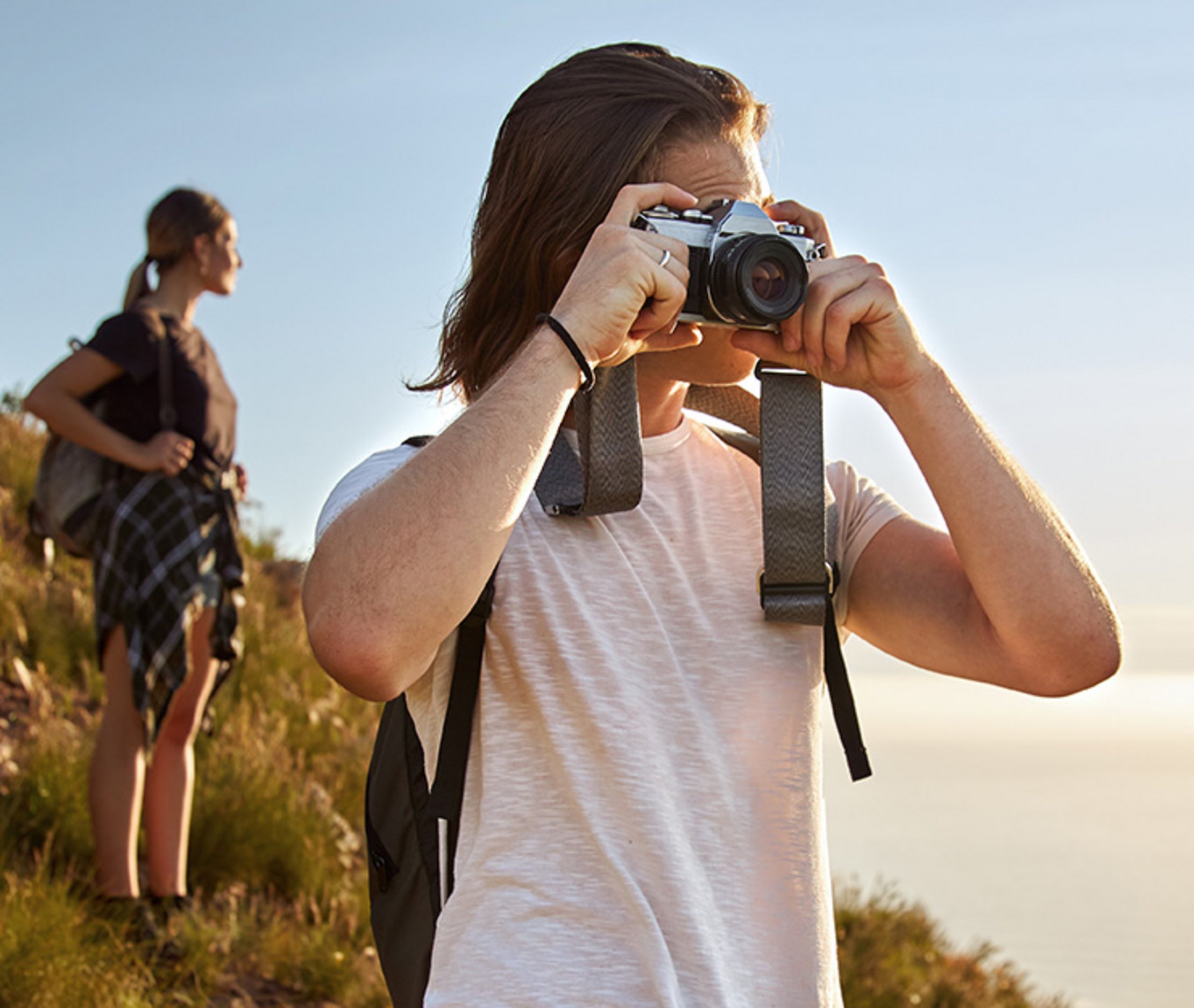Junger Mann fotografiert mit Kamera in der Natur beim Wandern, Frau im Hintergrund mit Rucksack – Outdoor-Abenteuer und Naturerlebnis bei Sonnenuntergang