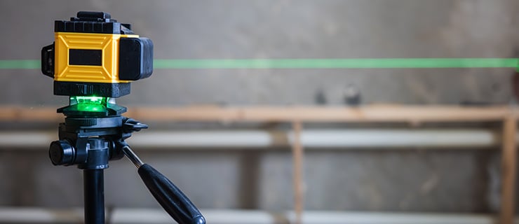 Lasergerät auf einem Stativ projiziert eine horizontale grüne Laserlinie auf einer Baustelle mit Betonwand und Holzgerüst im Hintergrund.
