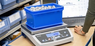 Eine blaue Kunststoffbox voller kleiner Metallteile steht auf einer digitalen Kern-Zählwaage, die das Ergebnis zeitgleich auf einem angeschlossenen Laptop-Bildschirm anzeigt.