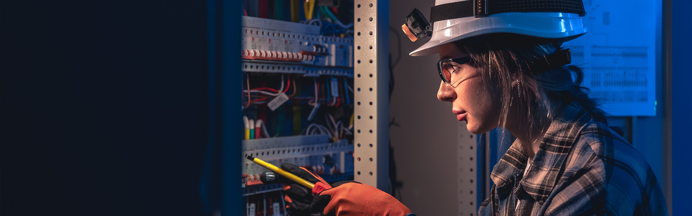 Elektrikerin mit Schutzhelm und Stirnlampe arbeitet mit Werkzeug an einem geöffneten Stromschaltschrank.