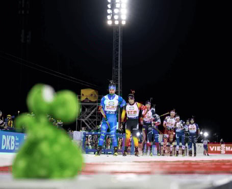 Eine Gruppe von Biathleten steht bei einem Nachtwettkampf in Östersund im Schnee, während im unscharfen Vordergrund eine grüne Figur zu sehen ist.