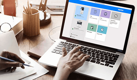 Eine Person tippt auf einem silbernen Laptop, der auf einem Holztisch steht und die Benutzeroberfläche der "Xerox Print and Scan Experience App" anzeigt, während die Person mit einem Stift in der anderen Hand Notizen in einem Notizbuch macht.