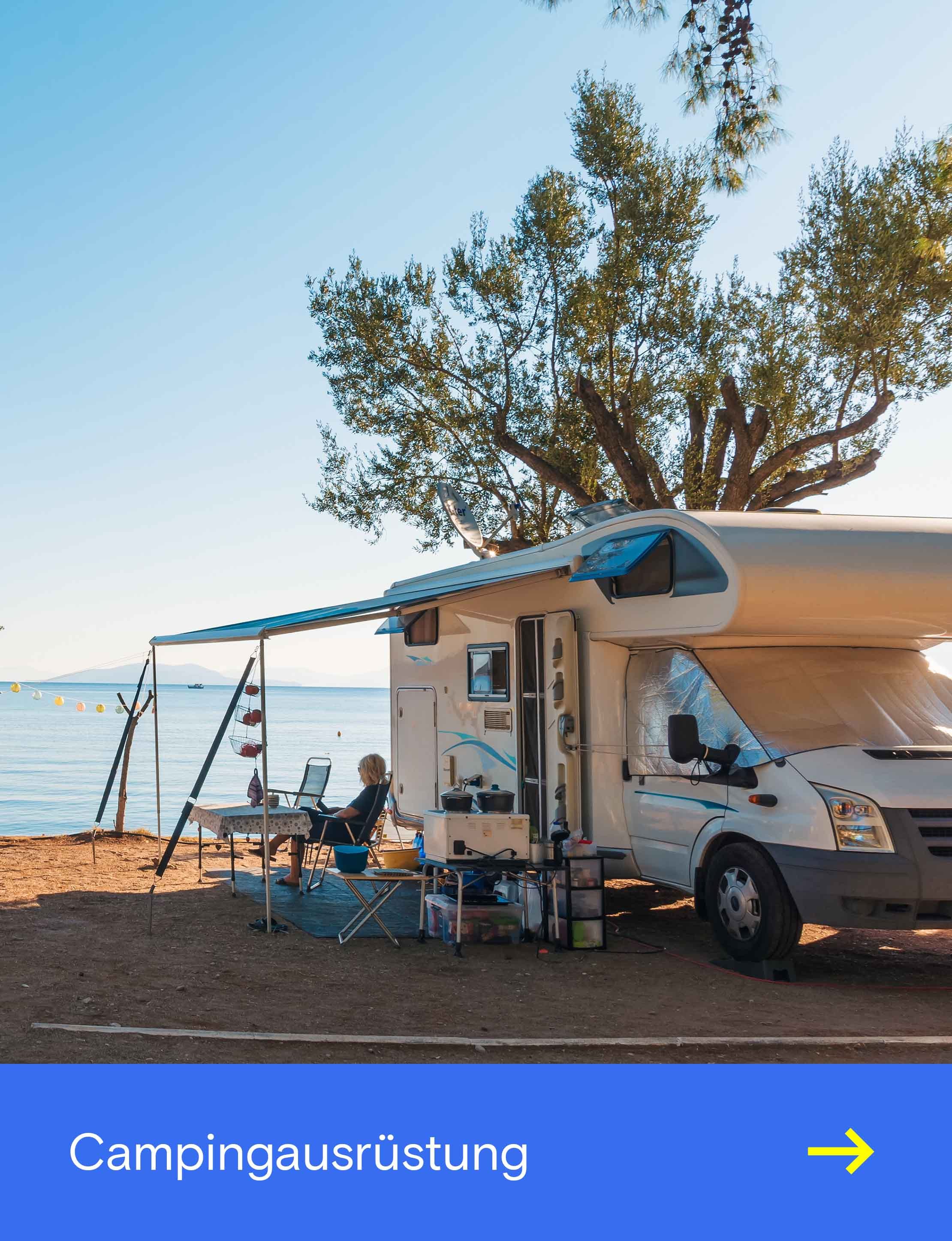 Ein Wohnmobil steht auf einem Campingplatz direkt am Meer, mit Markise, Stühlen und Blick auf das Wasser.