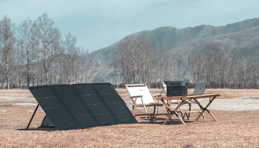 Solarpanel, Powerstation und Laptop auf einem Holztisch im Freien vor kahlen Bäumen und Bergen unter einem hellen Himmel. Ein weißer Stuhl steht daneben.