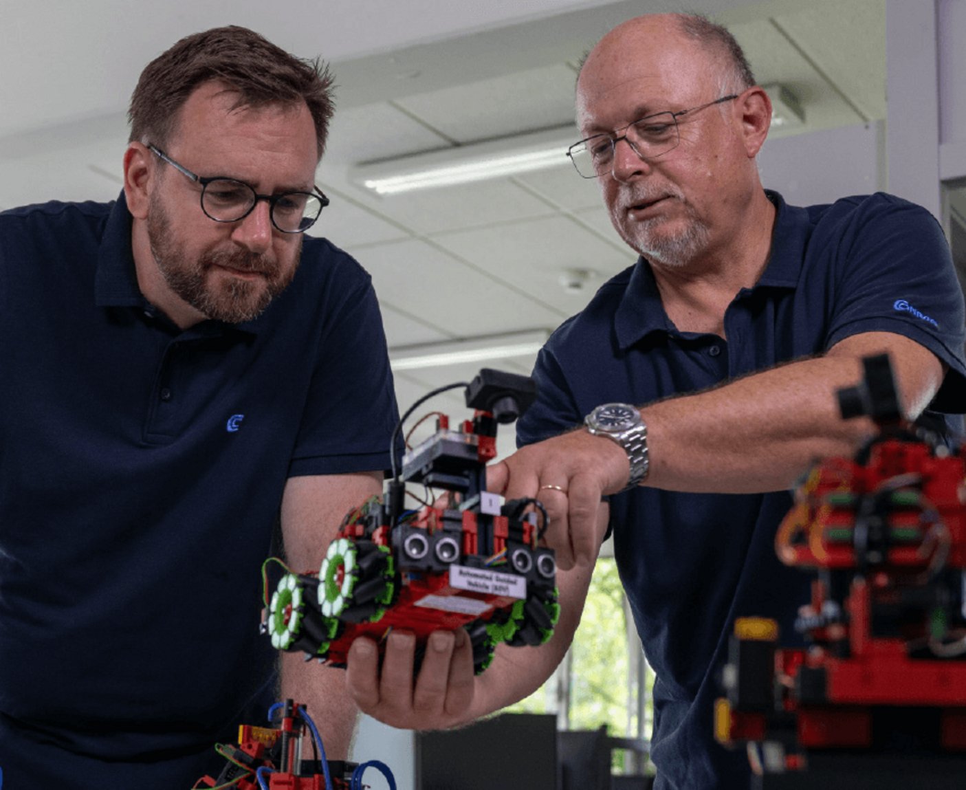 Wolfgang Lex und ein Mitarbeiter von Conrad, beide in dunklen Poloshirts begutachten konzentriert einen kleinen, aus roten und schwarzen Bauteilen konstruierten Roboter, des fahrerlosen Transportsystems aus dem Agile Production System.