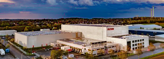 Eine Luftaufnahme zeigt das großflächige, moderne ELTEN-Logistikzentrum mit hellen Fassaden und Solaranlagen auf den Dächern unter einem dramatischen Abendhimmel.