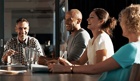 Vier lachende Personen sitzen an einem Besprechungstisch in einem hellen Büroraum, wobei die Person am rechten Bildrand unscharf ist und die anderen drei im Fokus sind.