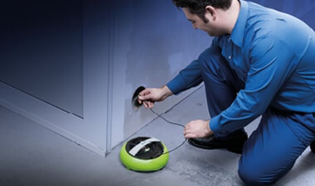 Ein Elektriker in blauer Arbeitskleidung kniet am Boden und führt ein Kabeleinzugsband aus einem grünen Gehäuse in eine Wandöffnung ein.