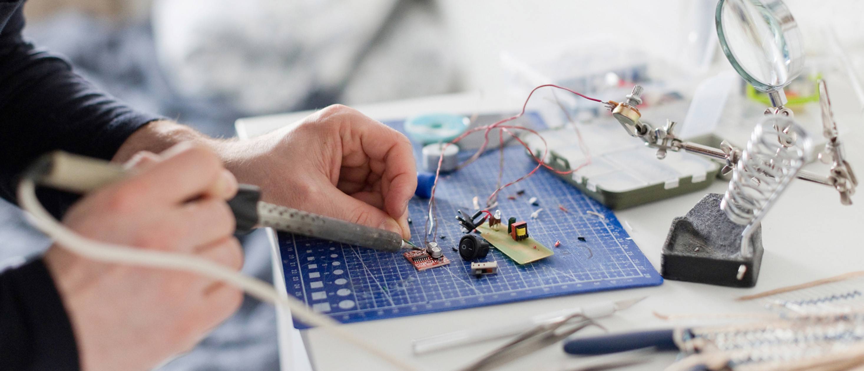 Nahaufnahme eines Lötvorgangs, bei dem eine Lötspitze auf einer Leiterplatte funkt und von Fingern präzise geführt wird.