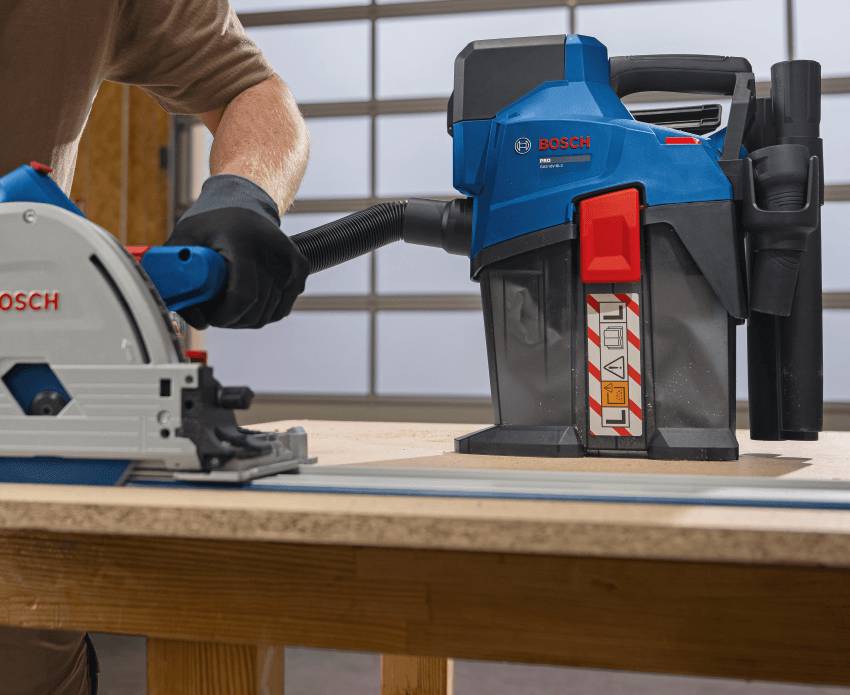 Eine Hand mit schwarzem Handschuh führt eine Bosch-Handkreissäge auf der Werkbank steht ein Akku-Staubsauger GAS 18 V-6LS der über ein Staubabsaugungsschlauch mit der Kreissäge verbunden ist und den Sägestaub absaugt.