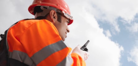 Ein Bauarbeiter mit orangefarbenem Schutzhelm und Warnweste blickt nach oben und spricht in ein Funkgerät vor einem bewölkten Himmel.