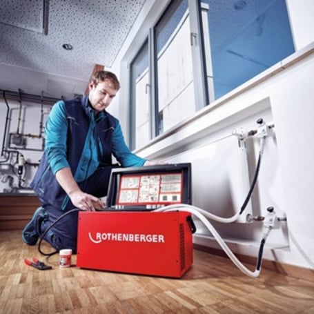 Ein HLK-Handwerker kniet neben einem roten Rothenberger Rohr-Einfriergerät auf einem Parkettboden und bedient die Klemmen, die an den Rohren eines Wandheizkörpers angebracht sind.